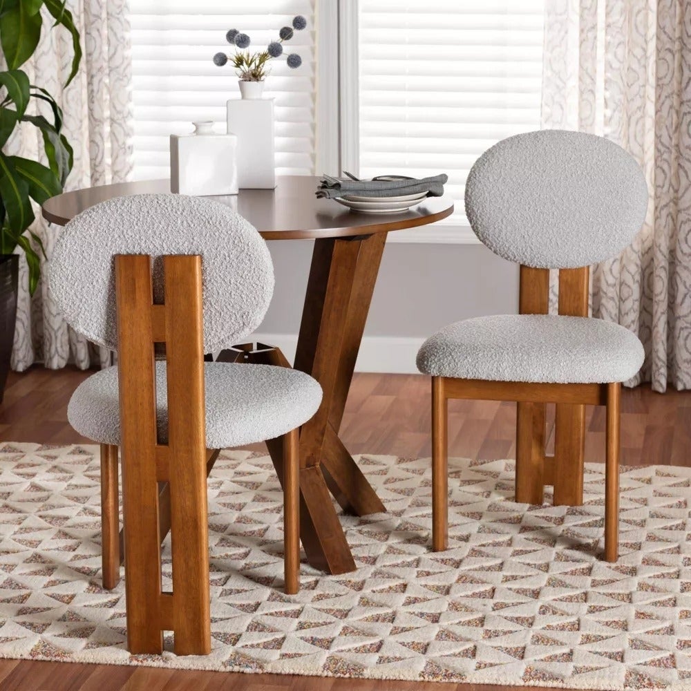 Meja bundar dengan kursi kayu modern berlapis kain bertekstur, diletakkan di atas karpet berpola geometris di sebuah ruangan dengan tirai putih dan tanaman pot