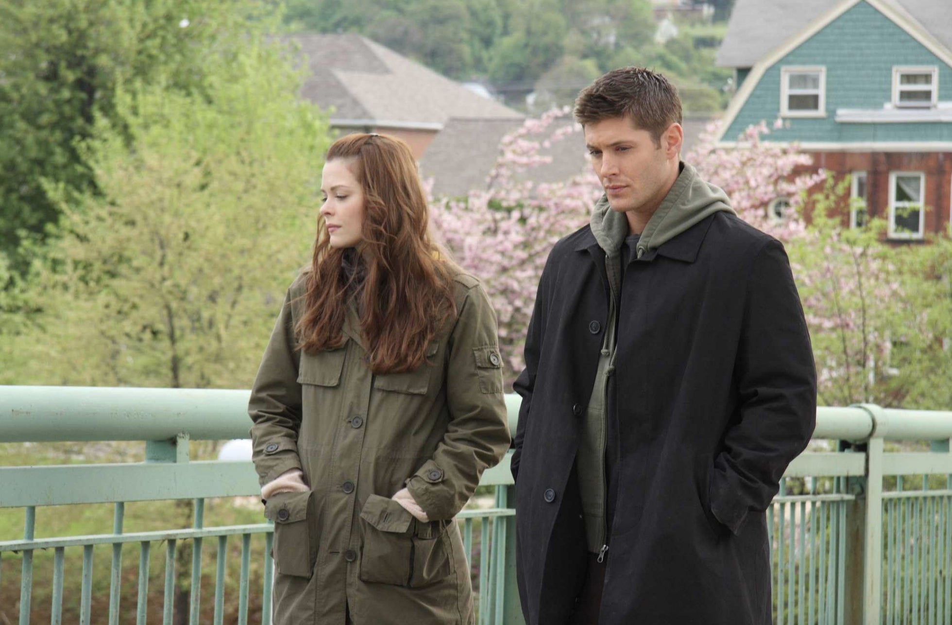 Two people in casual jackets stand on a bridge, looking pensive. Trees and houses are visible in the background