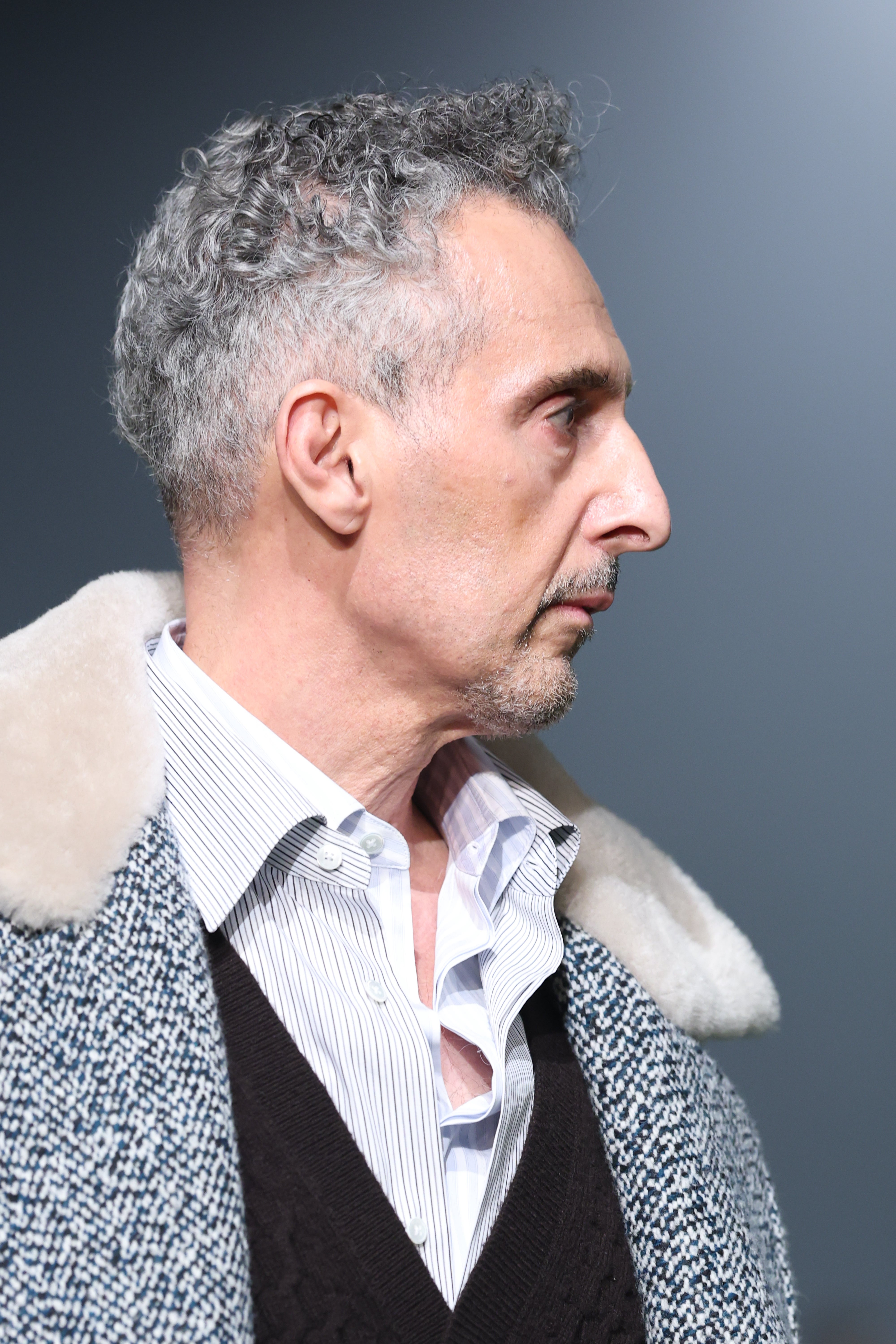 John Turturro with short, curly hair wearing a white collared shirt, black sweater, and a light coat with a plush collar, looking to the side