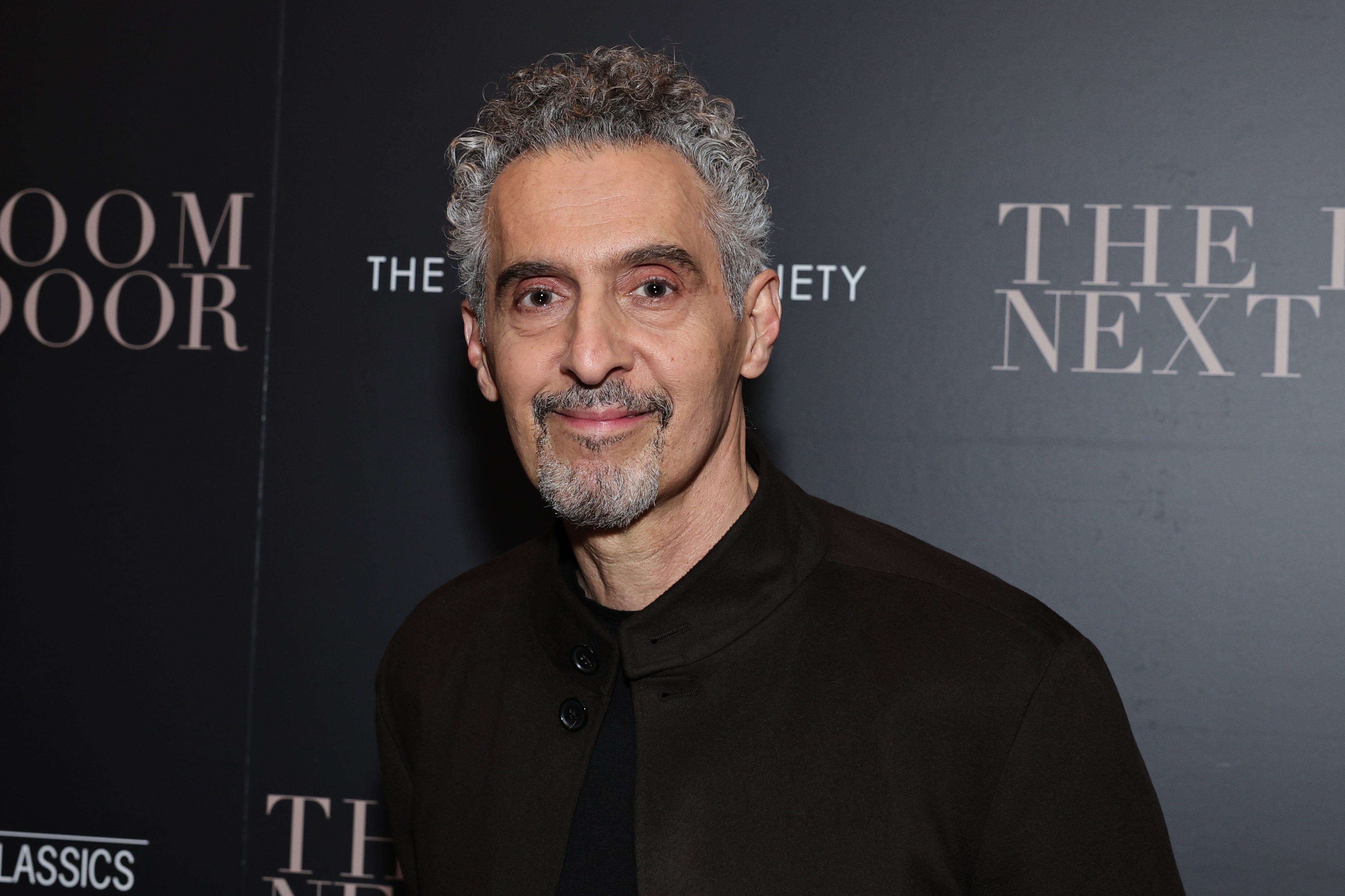 John Turturro in dark jacket at a media event backdrop for "The Room Next Door."