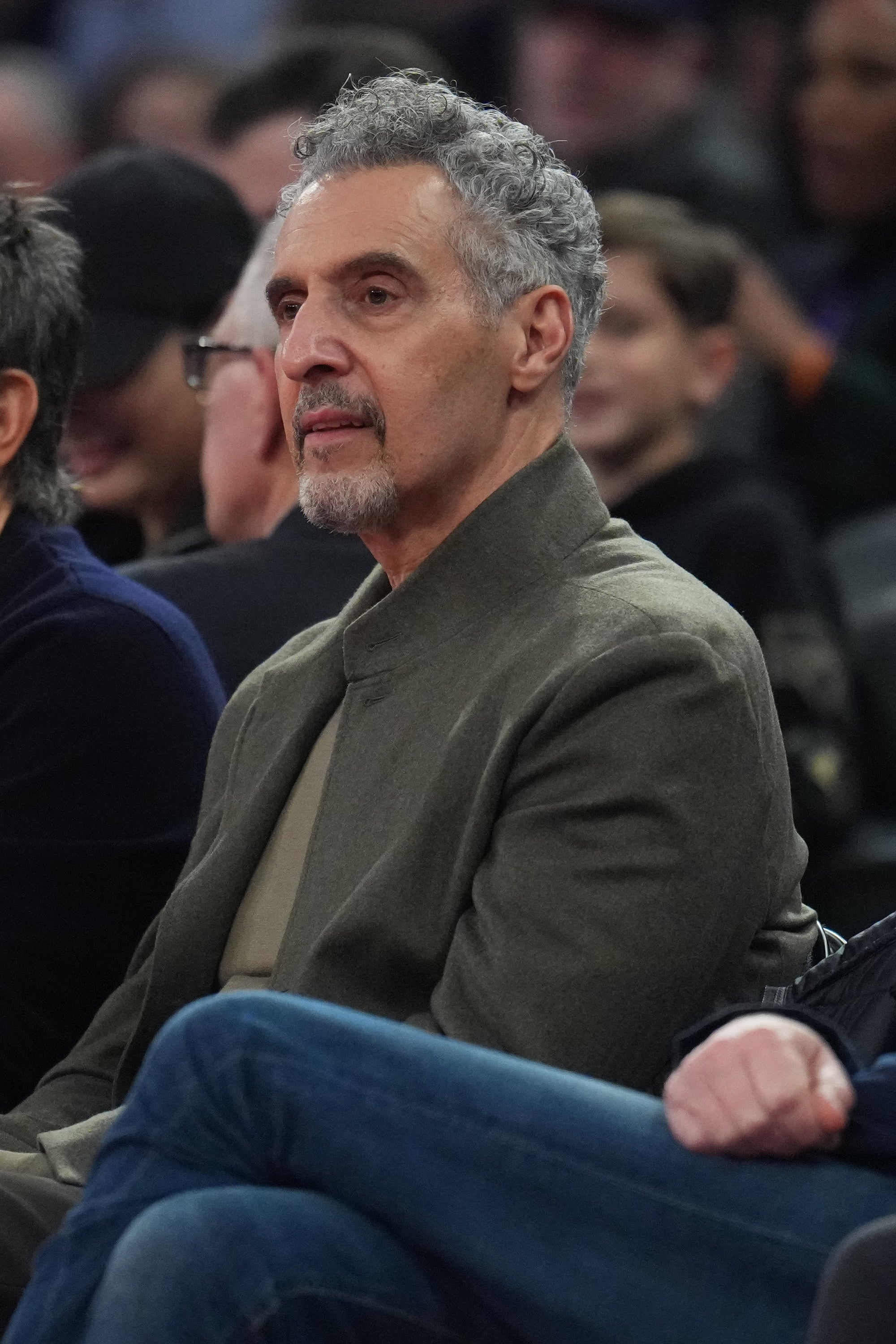 John Turturro sits at an event, wearing a simple, modern jacket