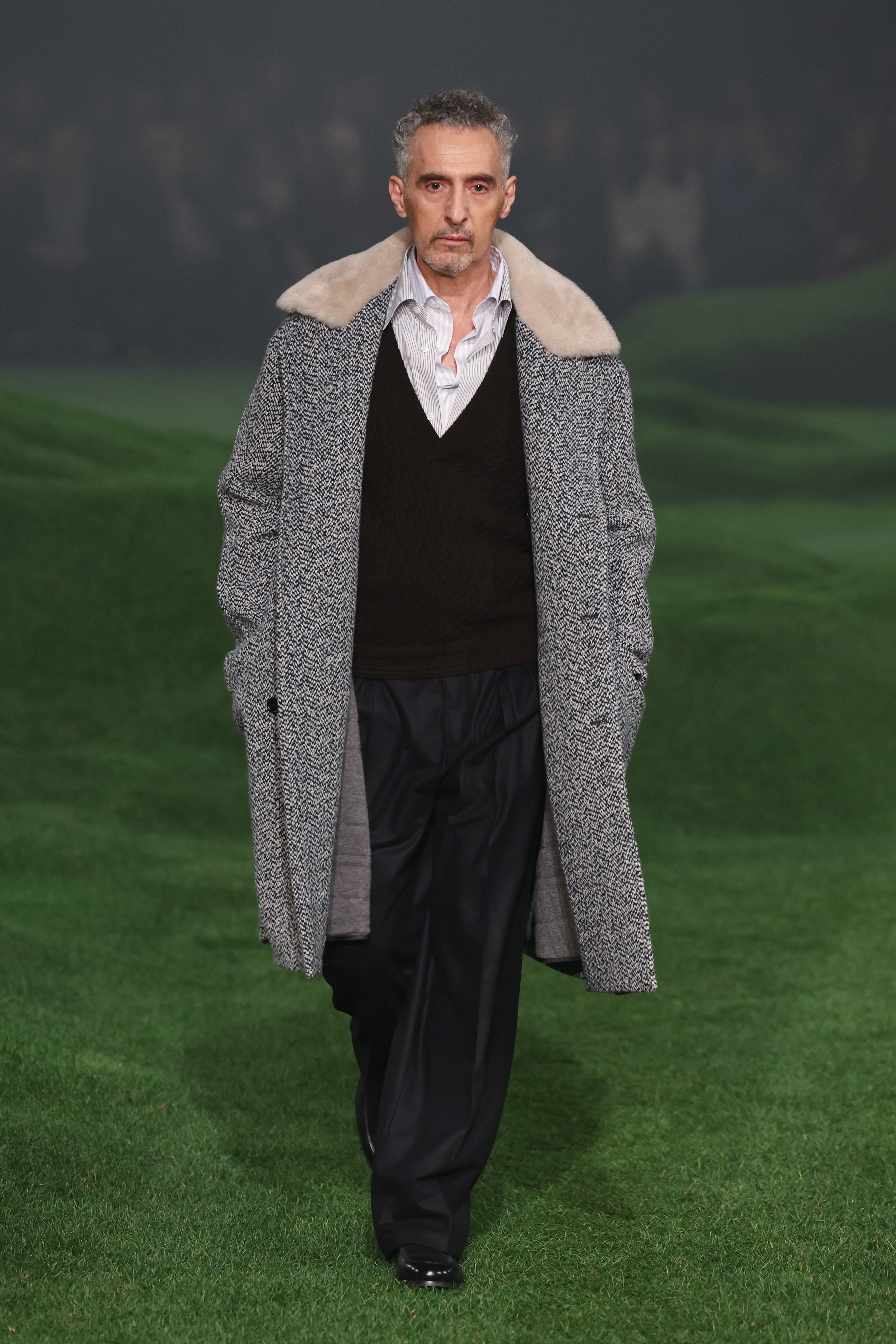 John Turturro walks on a grassy path wearing a textured coat with a fur collar, a black sweater over a white shirt, and black trousers
