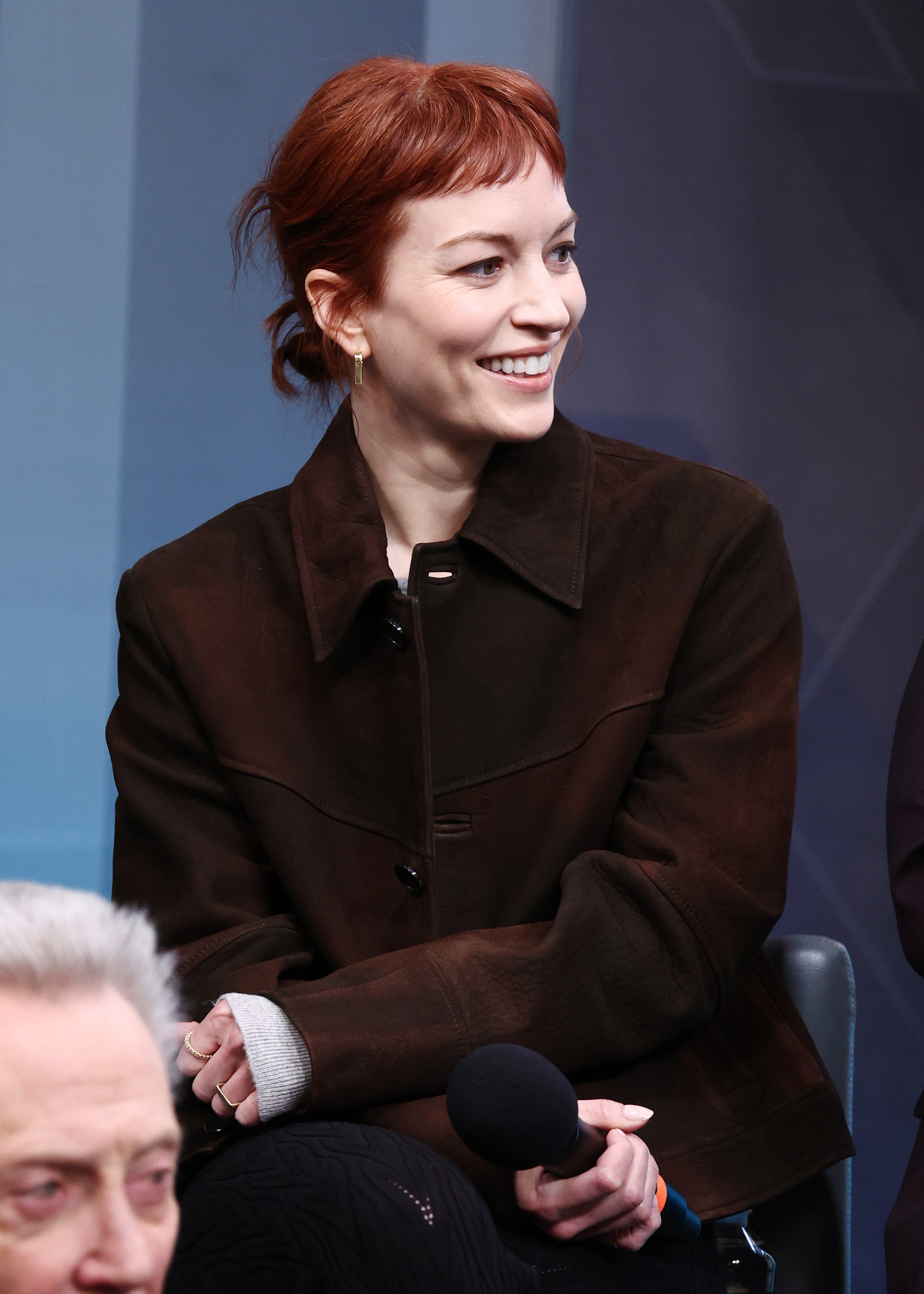 Britt Lower smiling in a brown jacket, seated with others during an interview or panel discussion