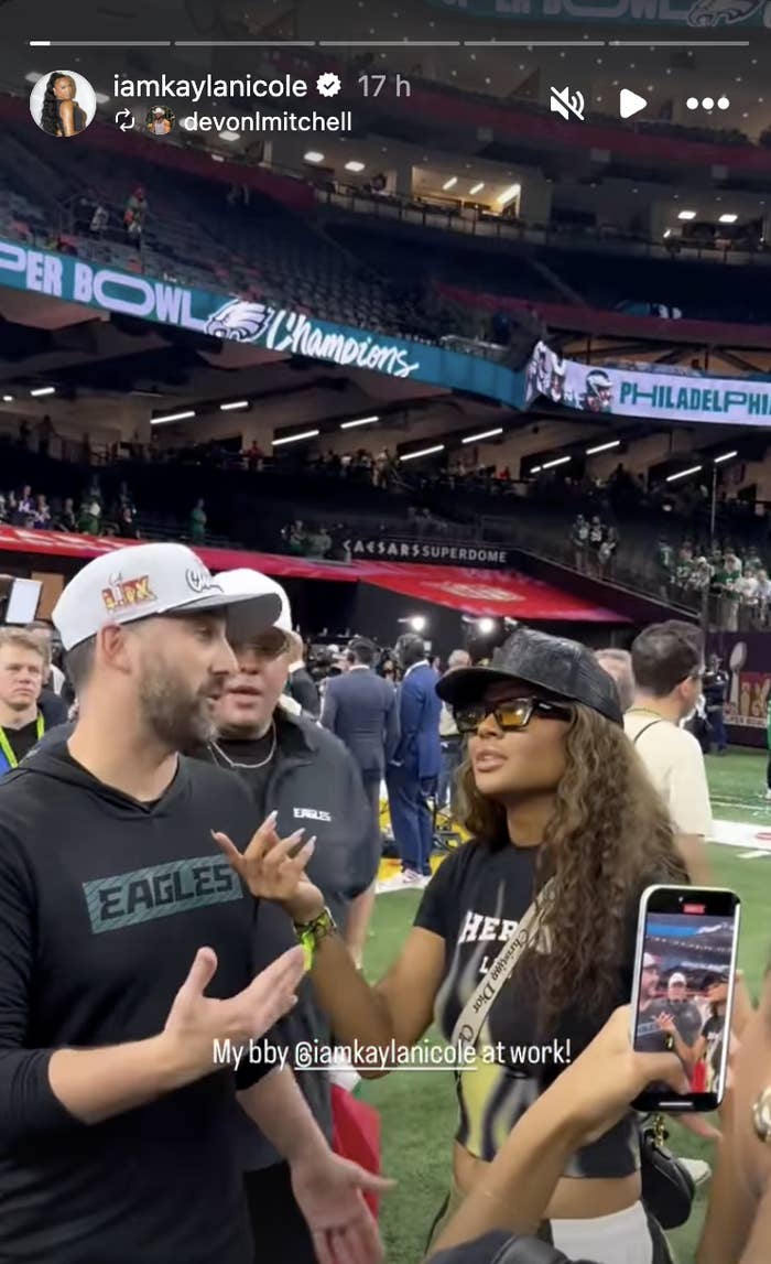 Person in Eagles shirt talking to woman in casual attire with a cap and sunglasses on the field. Background shows stadium and crowd