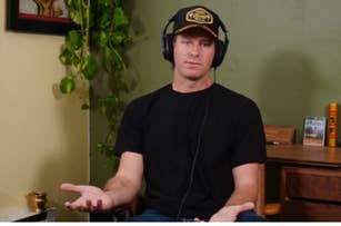 A person wearing headphones and a baseball cap sits indoors with a neutral expression, gesturing with open hands