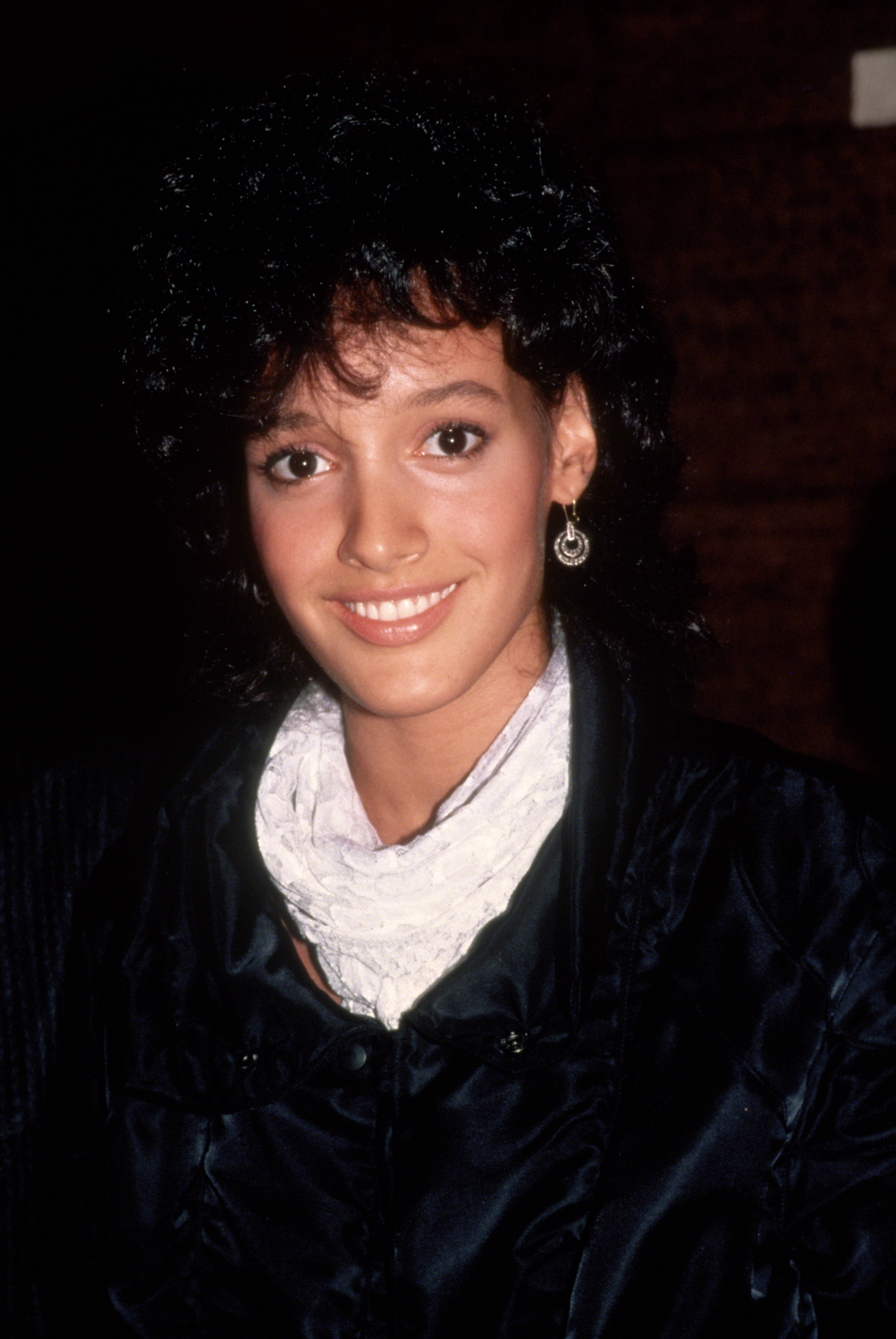 Person smiling, wearing a dark jacket and white scarf, with curly hair and hoop earrings