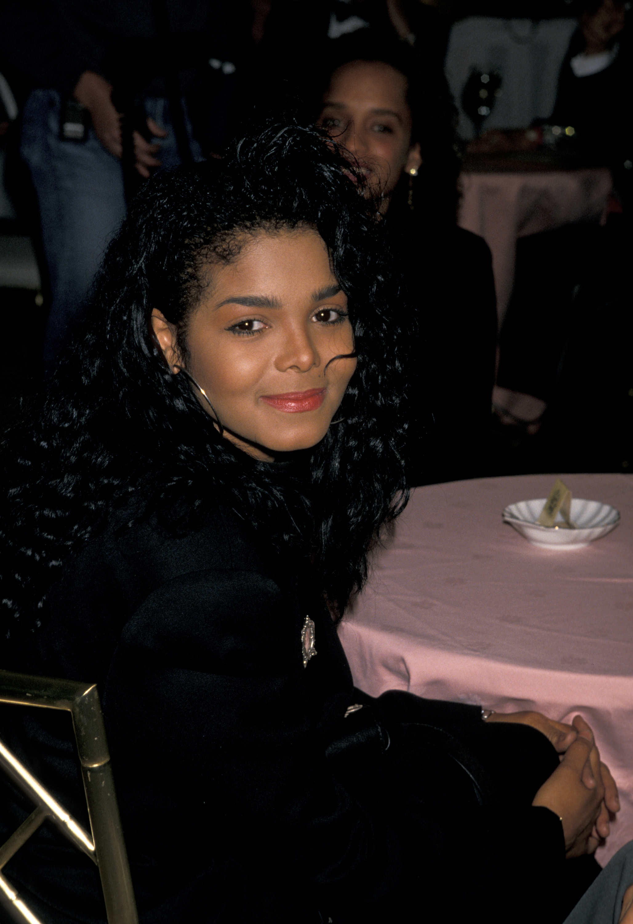 Person seated at a table, wearing a dark jacket with curly hair, smiling at the camera. Indoor setting with people in the background