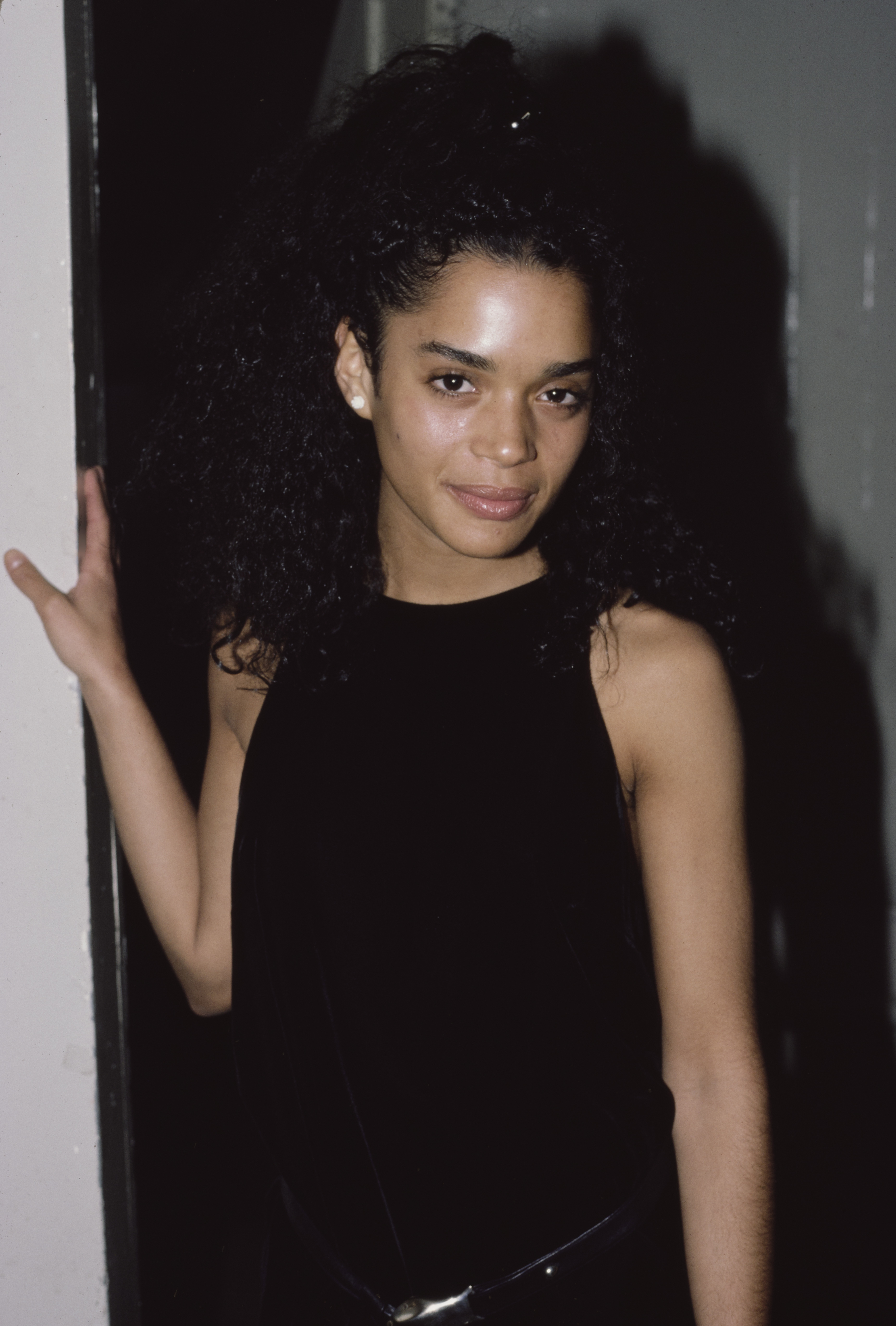 Person in elegant black sleeveless dress, posing indoors with a relaxed smile and curly hair styled up
