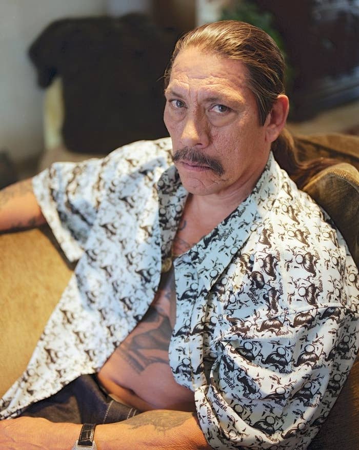Person seated with a serious expression, wearing a patterned open shirt