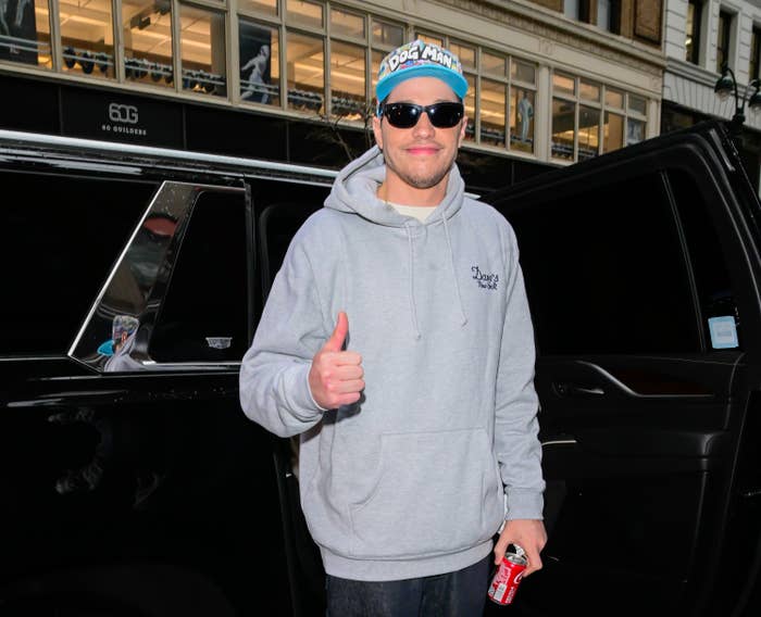 Person in a hoodie and sunglasses stands near a car, holding a soda can and giving a thumbs up