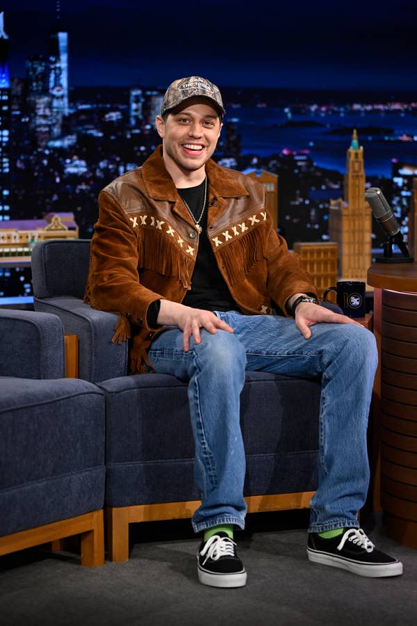 A person on a talk show set, wearing a camo cap, brown fringe jacket, black t-shirt, blue jeans, and sneakers, sitting and smiling