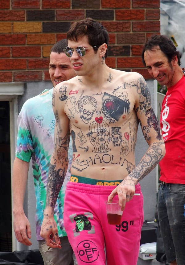 Person with numerous tattoos, wearing pink pants with a printed label, holding a cup. Two other people are nearby, smiling casually