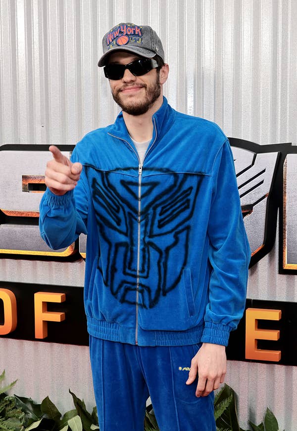 Person in blue velour tracksuit with graphic design, wearing a hat and sunglasses, pointing forward, stands at an event backdrop