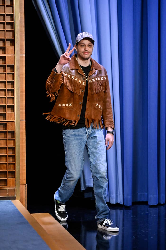 Person wearing a fringed jacket, jeans, sneakers, and a cap, making a gesture with fingers, on a stage with blue curtains in the background