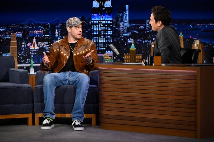 A person on a talk show wears a brown suede jacket and jeans, seated opposite the host at a desk with a night cityscape backdrop