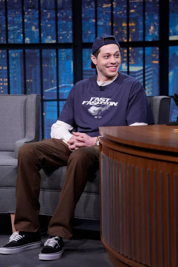 A person is seated on a talk show set wearing a dark T-shirt over a long-sleeve shirt, brown pants, and a cap turned backward, smiling