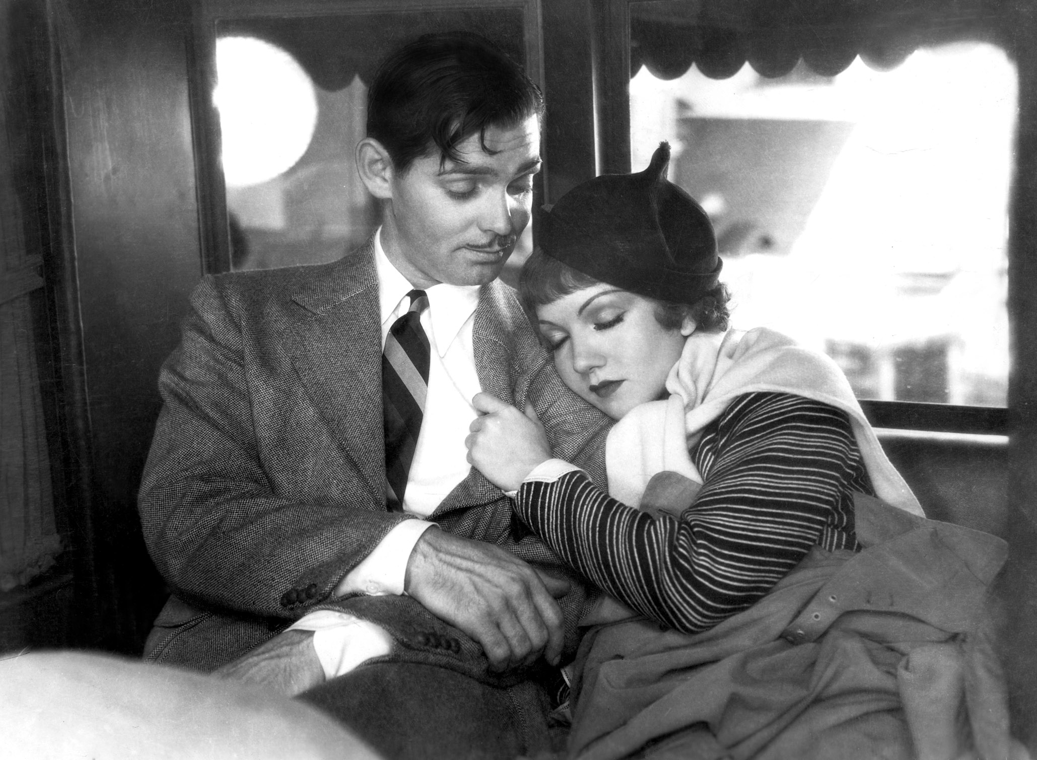 A classic film scene with a man in a suit and a woman in a striped dress and hat, sharing a tender moment together