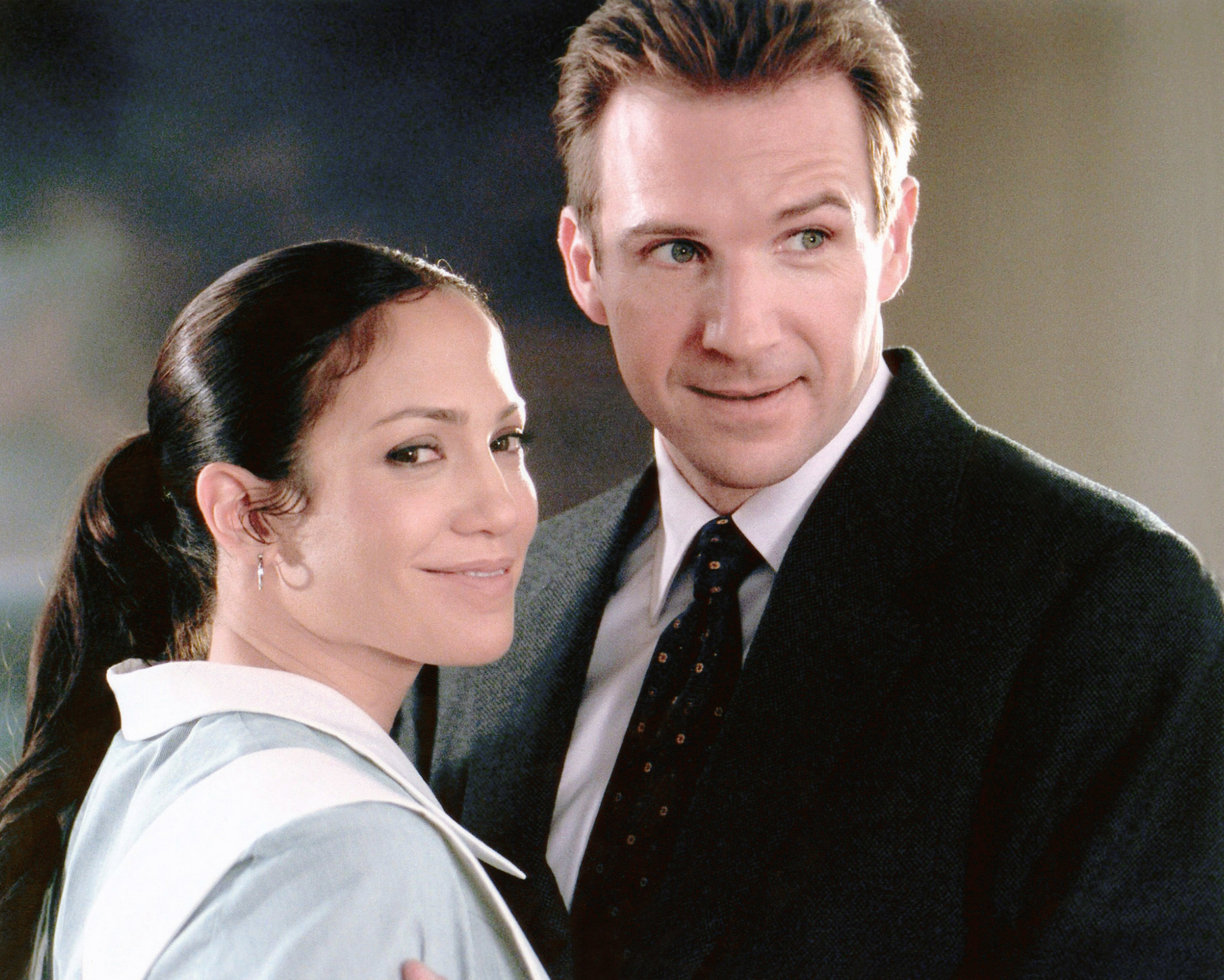 Two individuals in a formal setting, one in a collared outfit and the other in a suit and tie, smiling and looking toward the camera