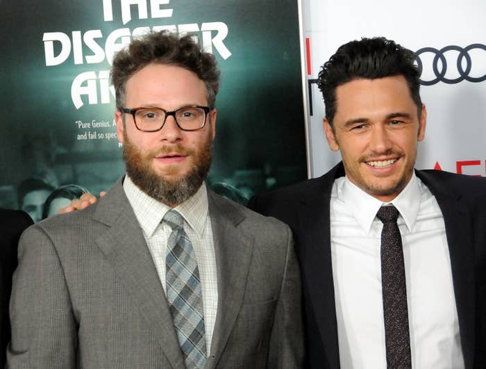 Two individuals in suits at a movie premiere event. One wears glasses and a patterned tie, the other has a solid tie and is smiling