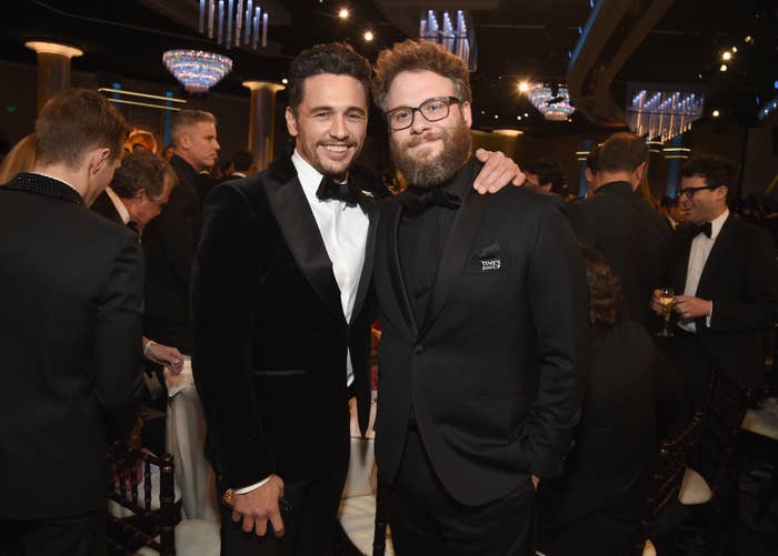 Two celebrities wearing tuxedos pose together at a formal event, surrounded by other elegantly dressed attendees