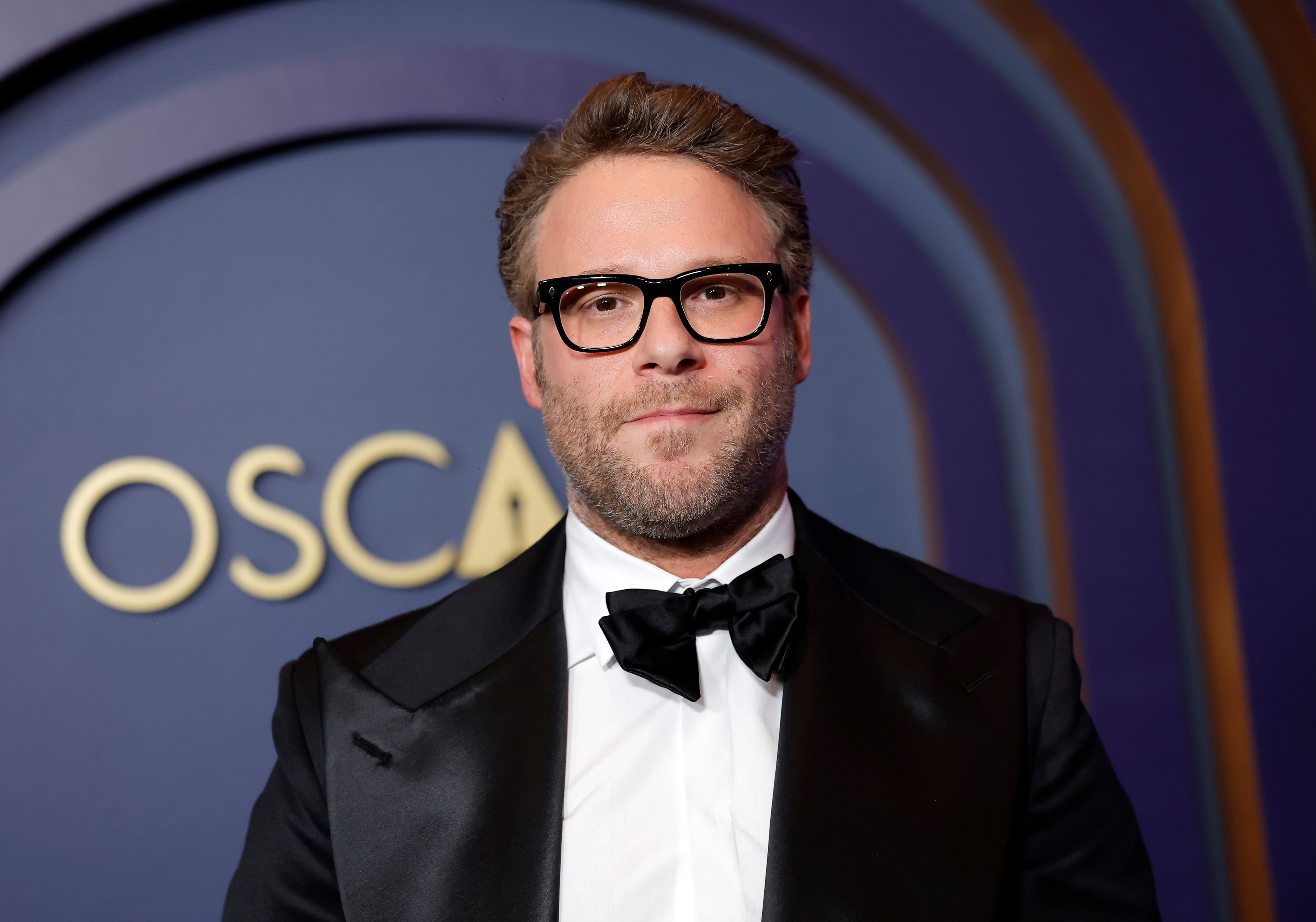Man in formal black tuxedo with glasses stands at an event with an "Oscar" sign in the background