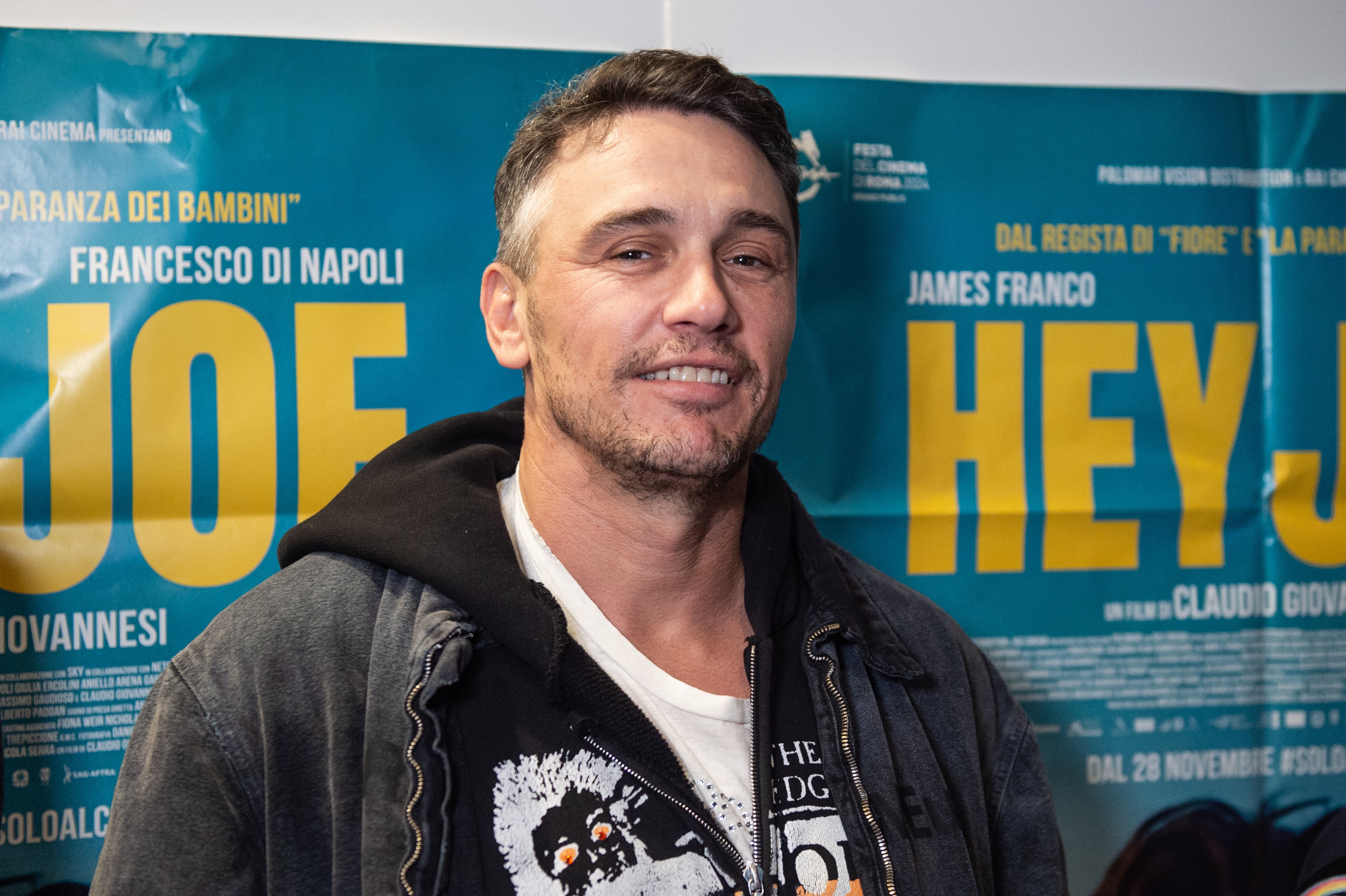Person in casual jacket at a film premiere, standing in front of a movie poster featuring large text "JOE" and "HEY JOE."