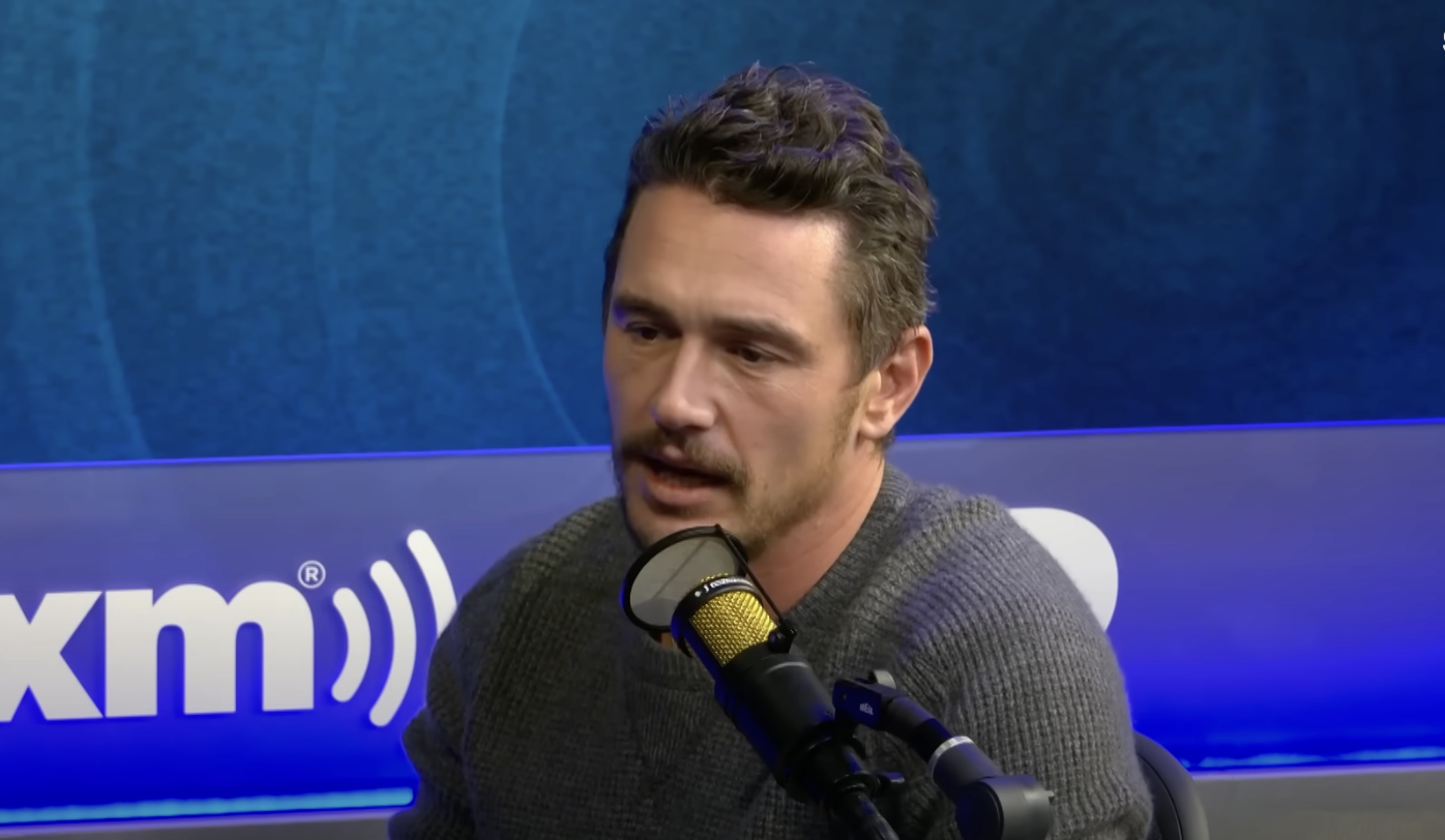 A person speaking into a microphone in a studio setting, wearing a textured sweater