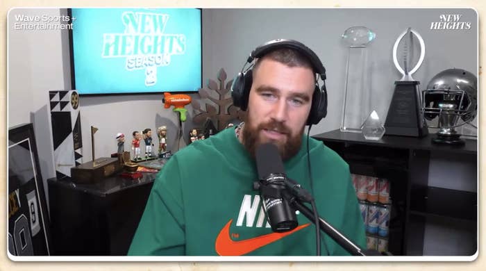 Traivs Kelce recording a podcast in a studio, wearing a sweatshirt and headphones, sitting in front of a microphone and sports memorabilia