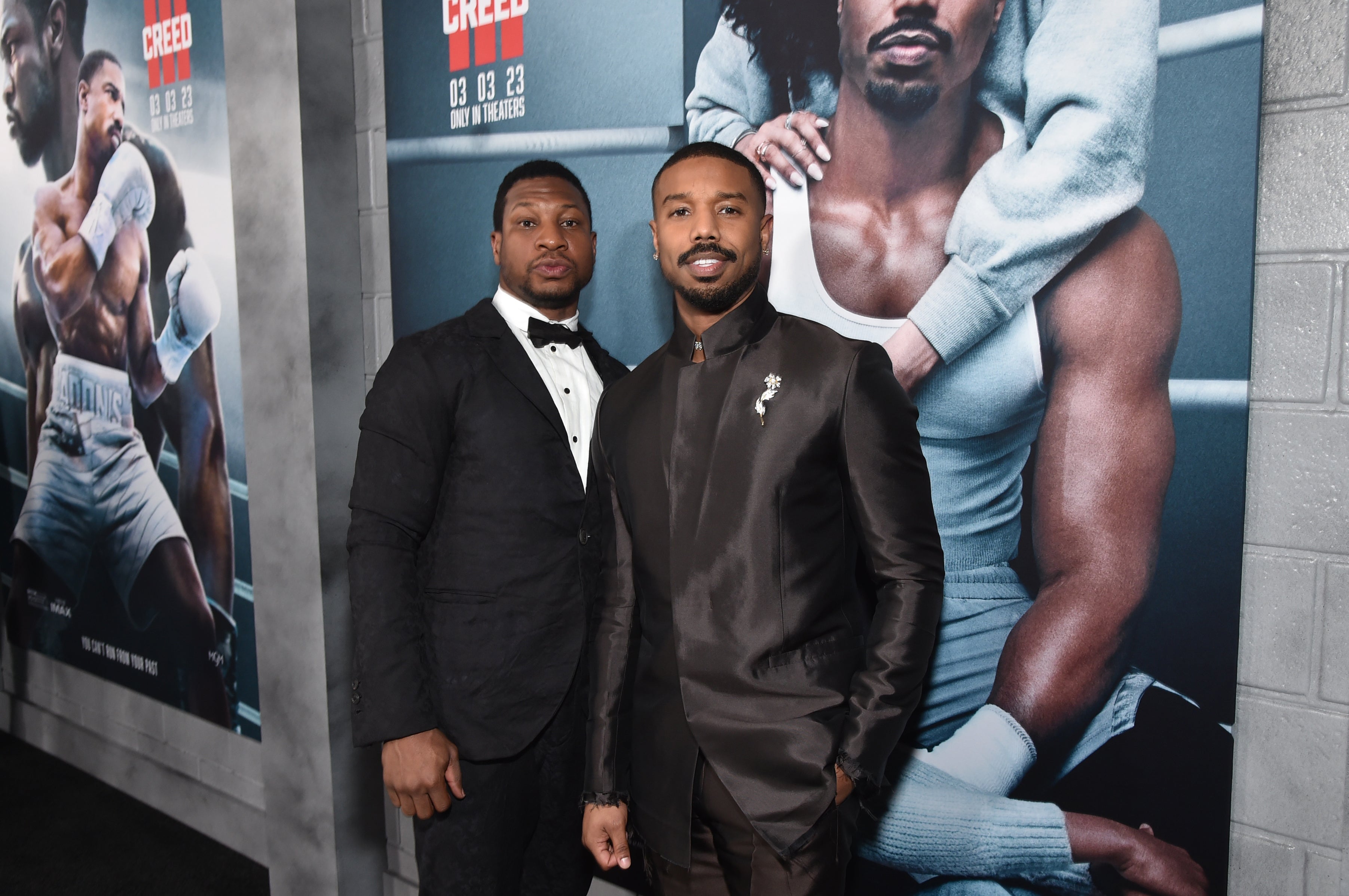 Michael B. Jordan and Jonathan Majors in formal suits posing at a movie premiere, standing in front of a "Creed III" poster