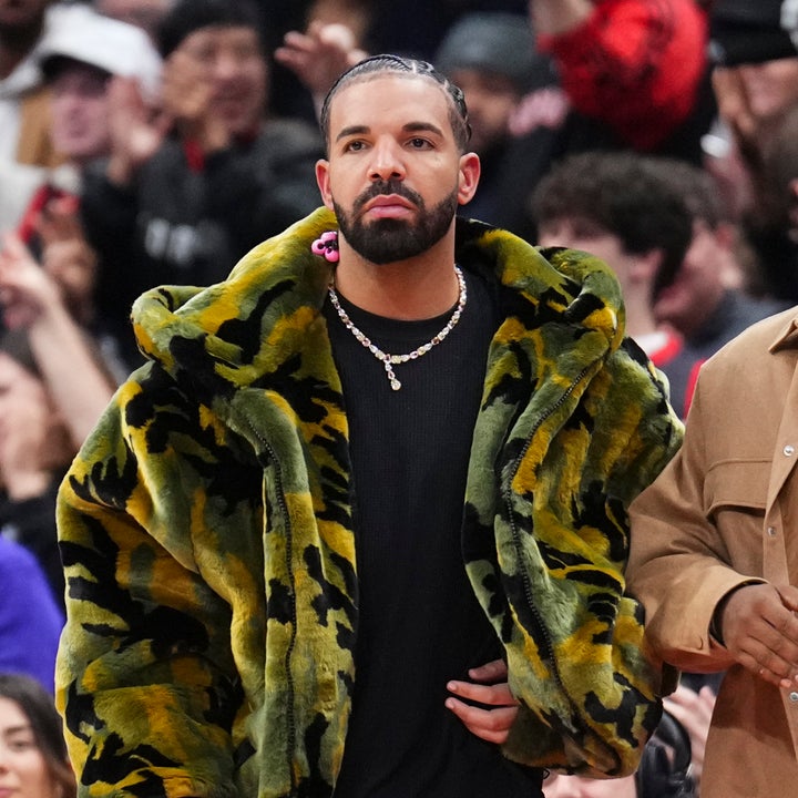 Drake in a bold, oversized fur coat stands at a public event, surrounded by a crowd