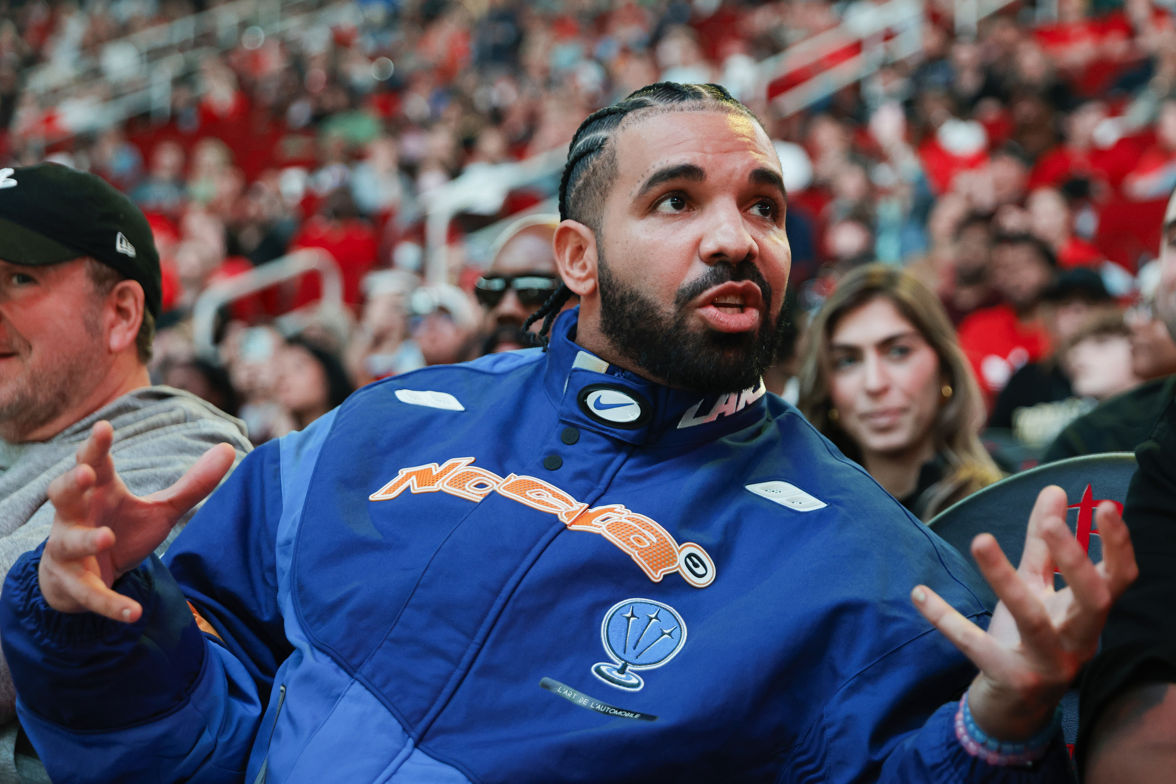 Drake in a sporty jacket sits in a crowded venue, gesturing with hands while engaged in conversation