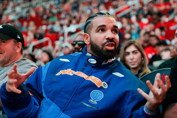 Drake in a sporty jacket sits in a crowded venue, gesturing with hands while engaged in conversation