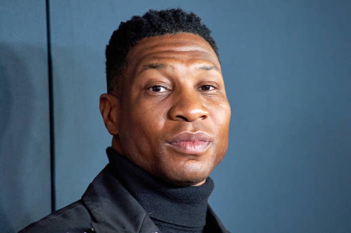 Jonathan Majors wearing a black turtleneck and jacket, looking at the camera against a plain background