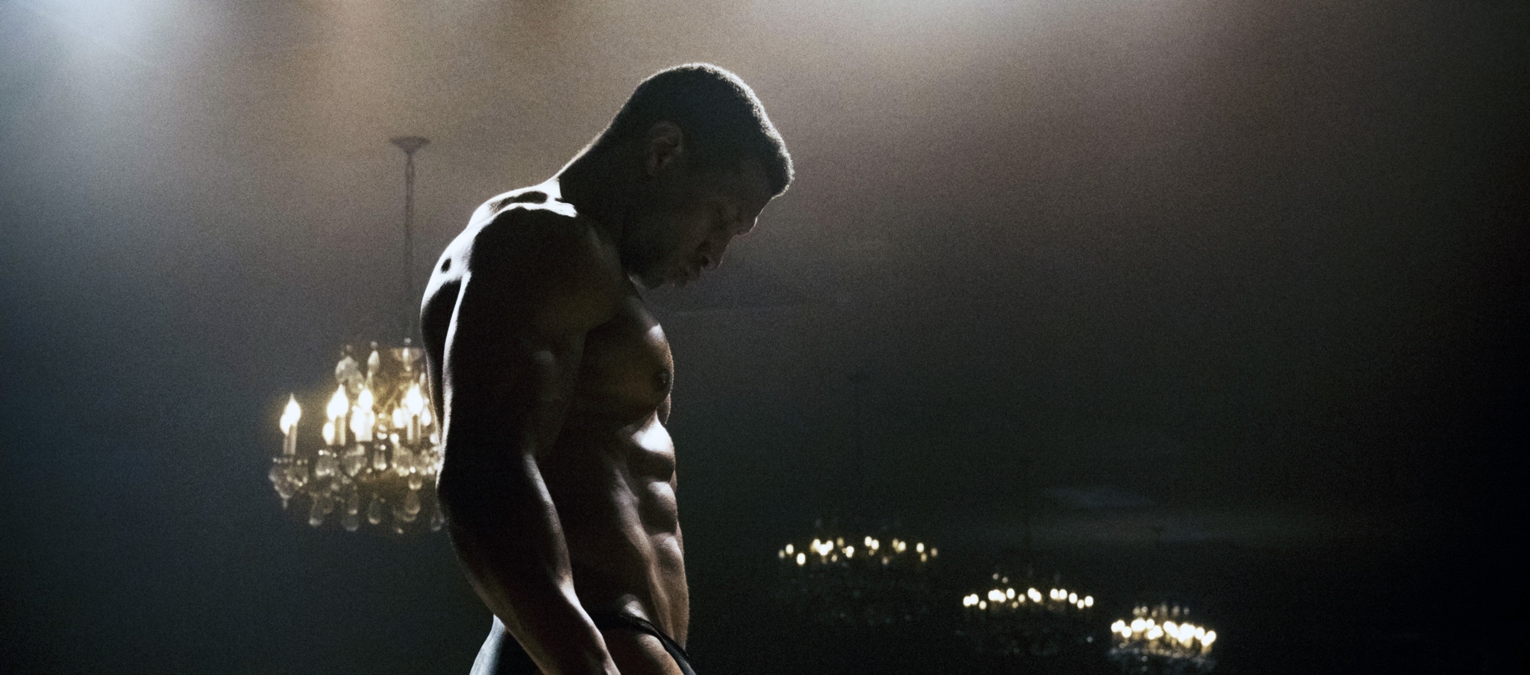 Jonathan Majors stands shirtless with his head down, surrounded by chandeliers in a dimly lit room