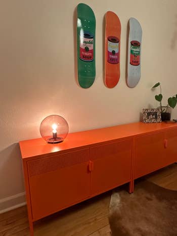 Room with orangish sideboard, circular lamp, and wall-mounted skateboards featuring crockery tin designs. Stylish location decor