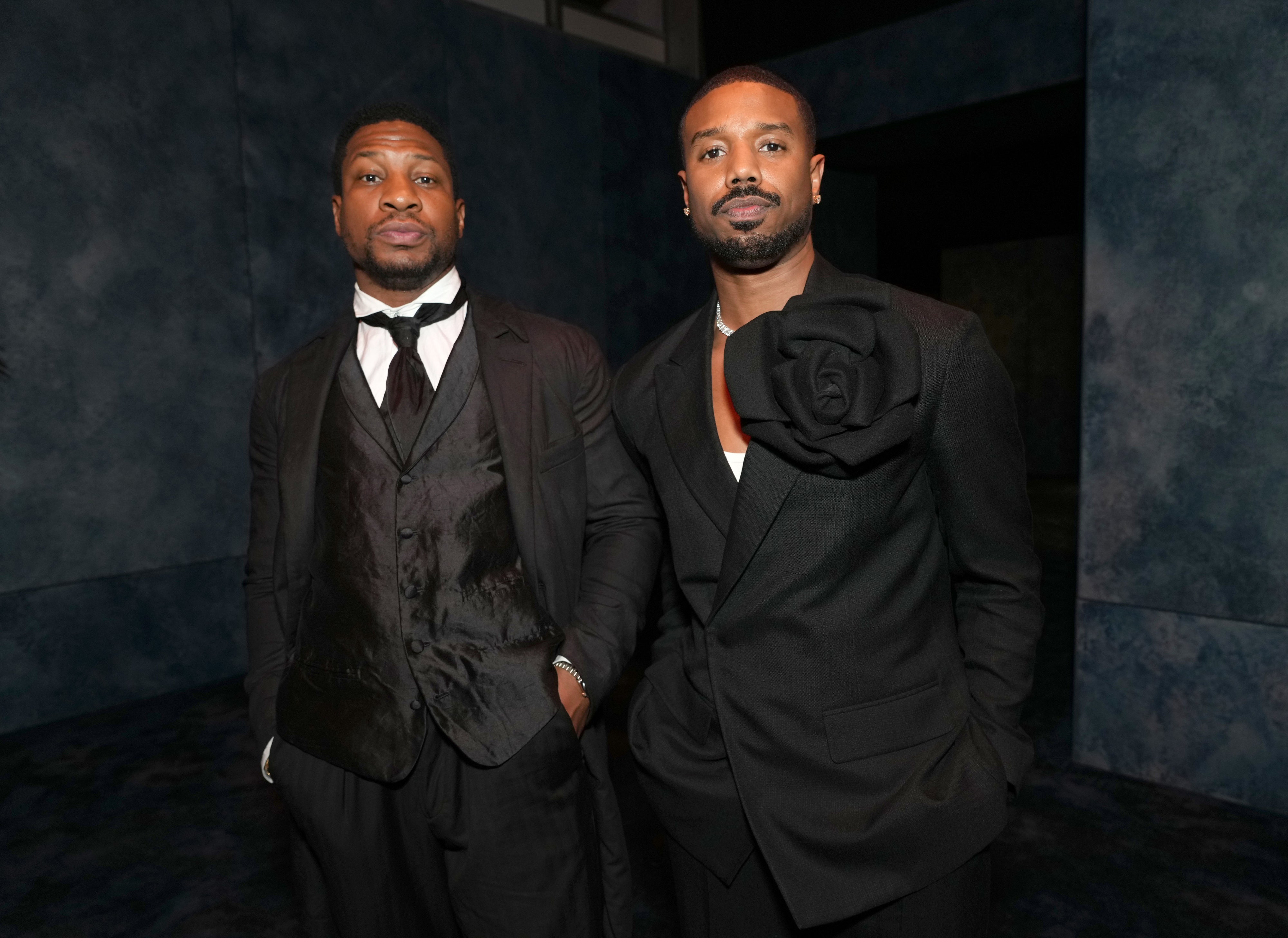 Jonathan Majors and Michael B. Jordan in formal suits pose together at an event. One suit features a large floral detail on the lapel