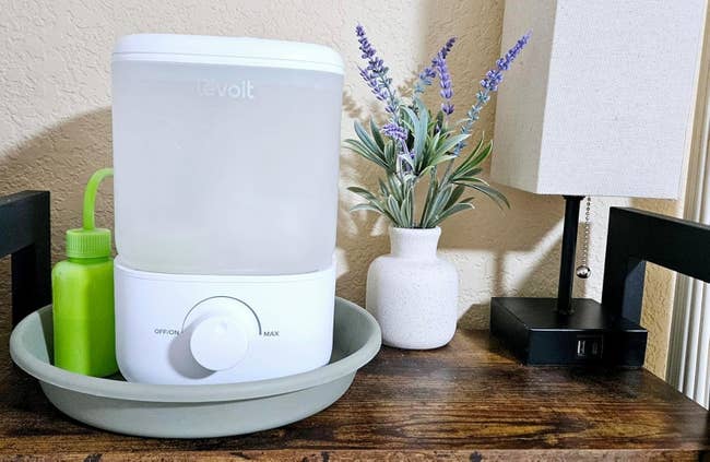 Humidifier on a tray next to a small green bottle, vase with lavender, and a table lamp
