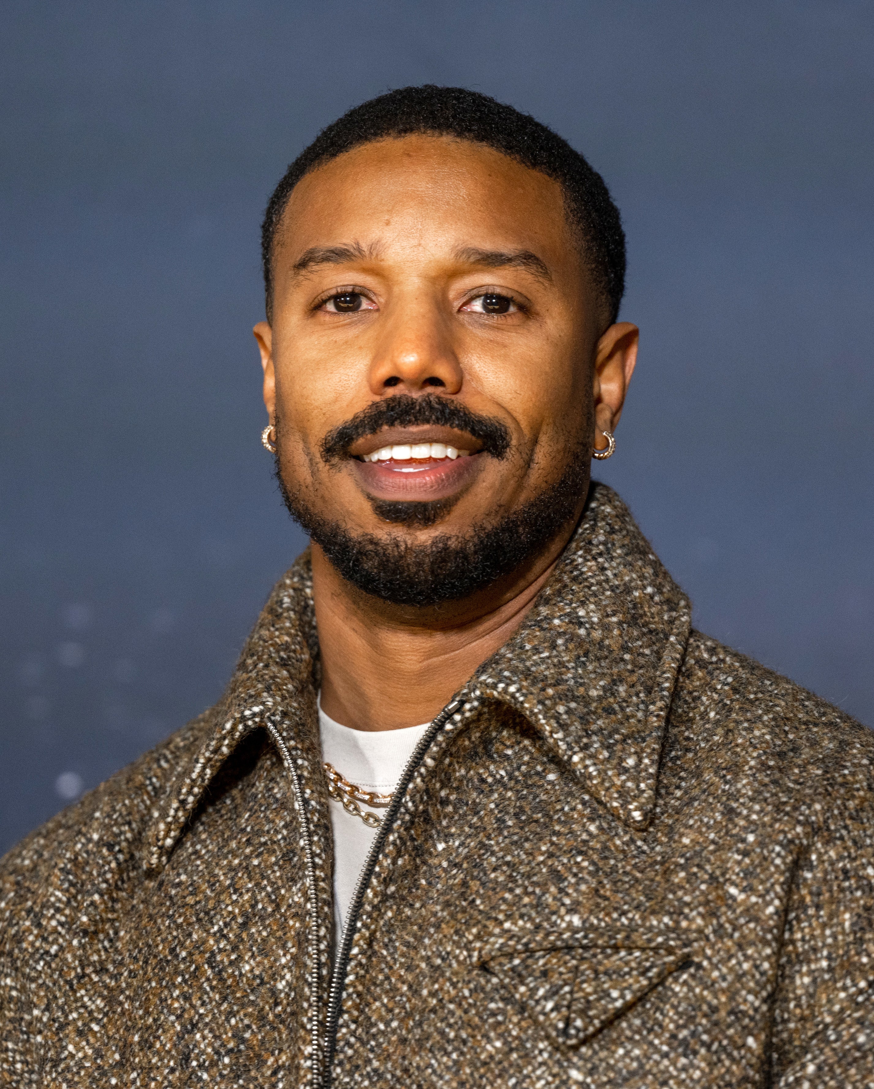 Michael B. Jordan smiling, wearing a textured jacket and T-shirt, against a plain background