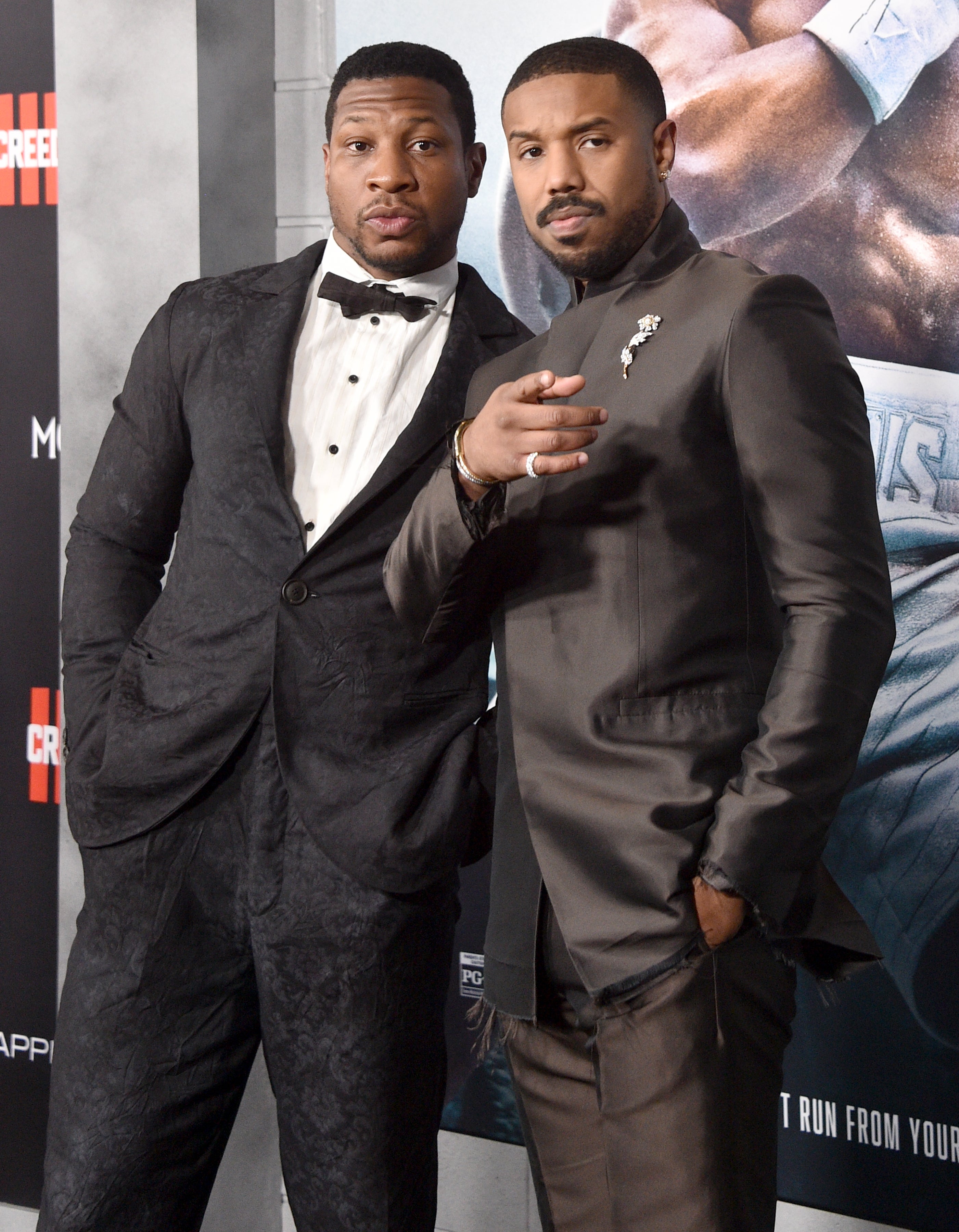 Jonathan Majors and Michael B. Jordan on a red carpet, one in a velvet tuxedo and the other in a sleek, modern suit with floral embellishments, pose for the camera