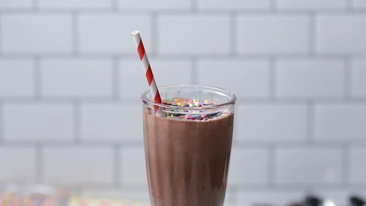 A chocolate milkshake topped with colorful sprinkles and a striped straw sits in a glass with a tiled background