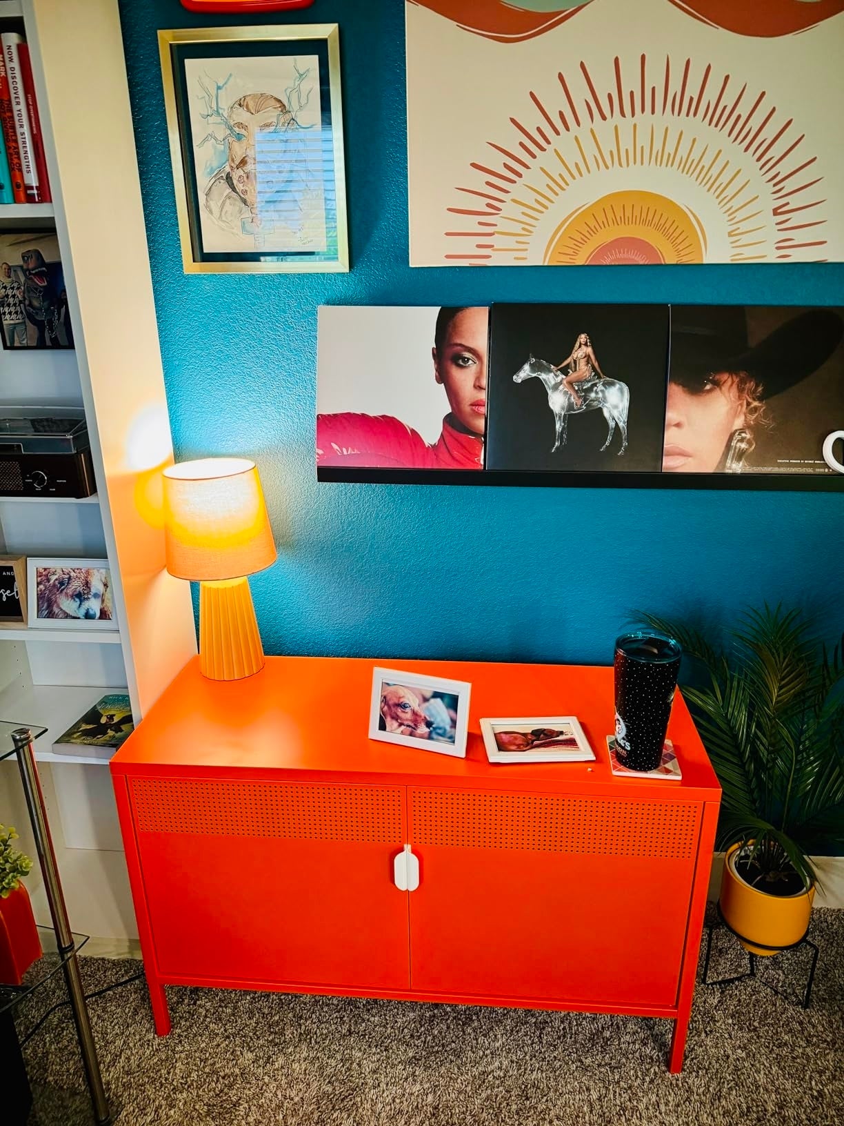 Room with a vibrant orangish  cabinet, decorative artwork, and a support  of books. Two framed photos remainder  connected  the cabinet, adjacent  to a lamp and a drink