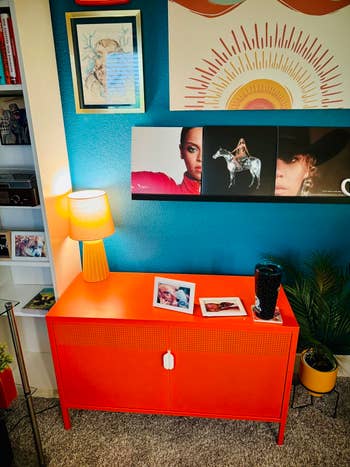 Room with a vibrant orangish cabinet, decorative artwork, and a support of books. Two framed photos remainder connected the cabinet, adjacent to a lamp and a drink