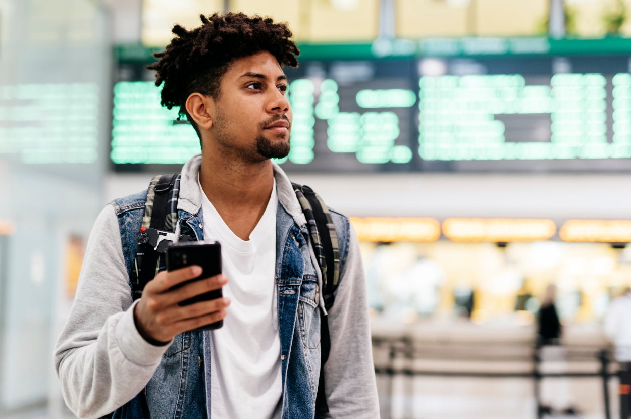 Person at airport, casually dressed in a denim jacket with a backpack, holding a phone. Departure board with blurred details in the background
