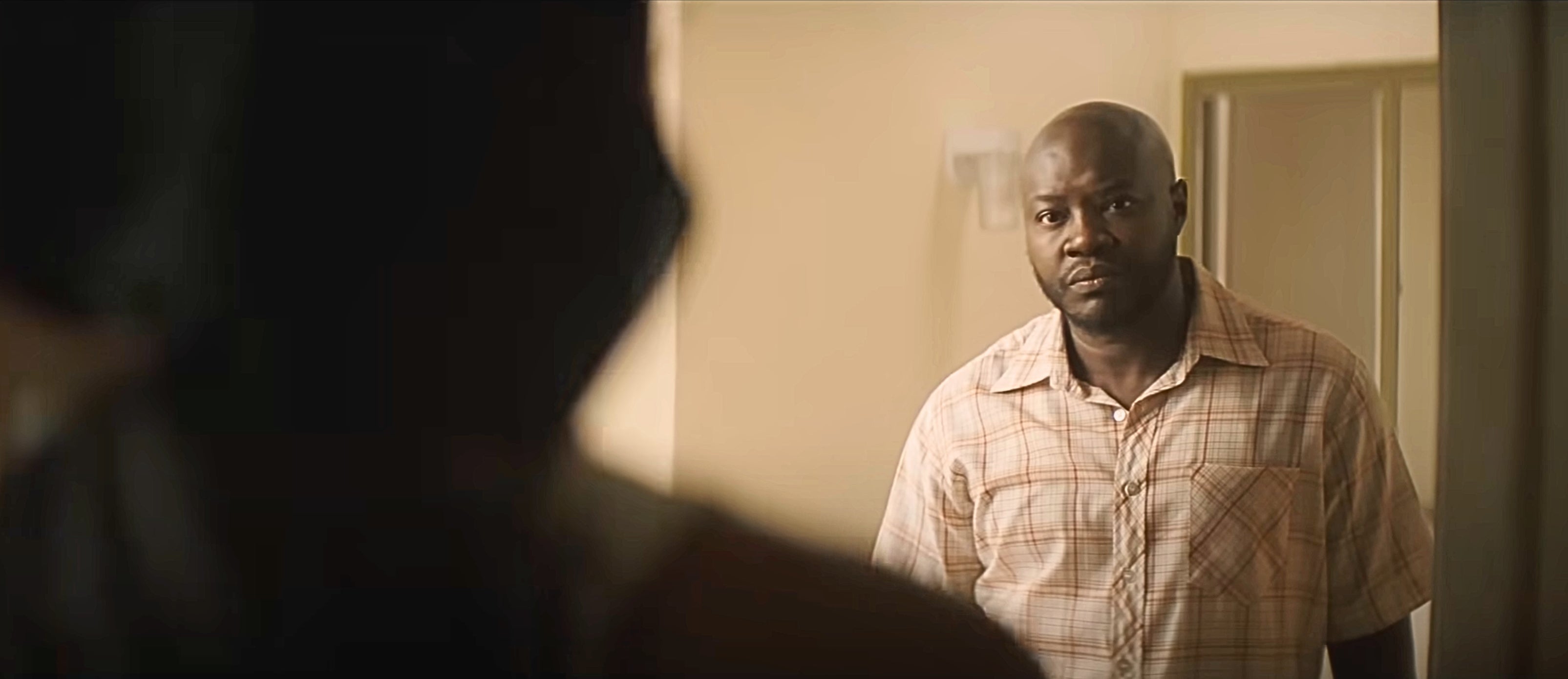 A man in a plaid shirt looks into a dimly lit room with a neutral expression