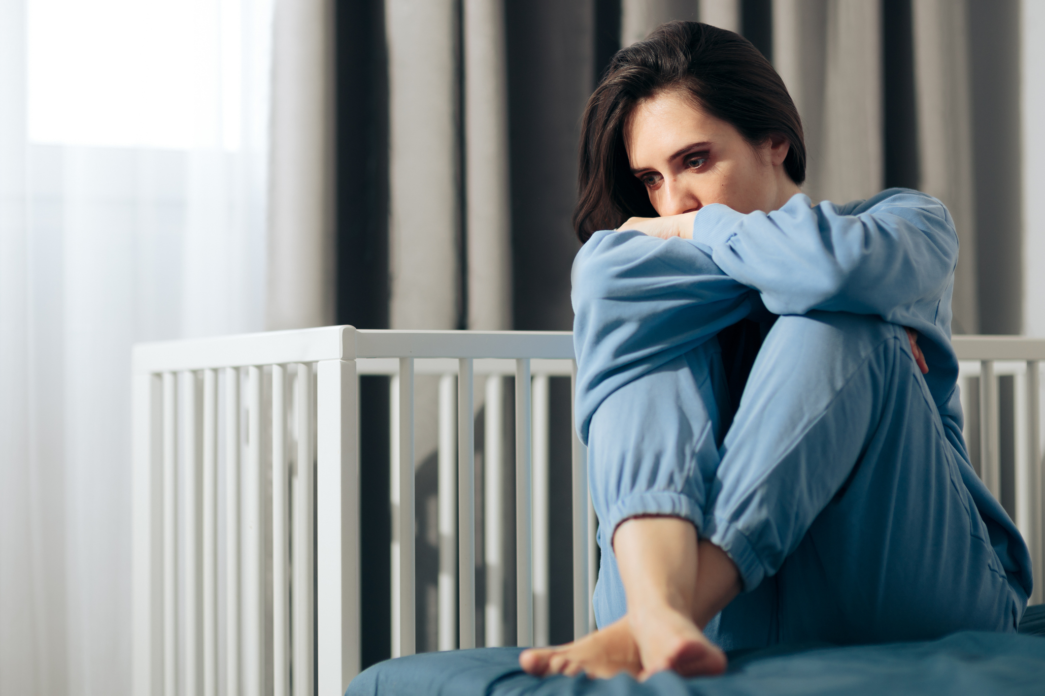 Person som sitter pensivt på en seng, iført komfortabelt loungewear, armene pakket rundt knærne, ved siden av en hvit barneseng i et mykt opplyst rom