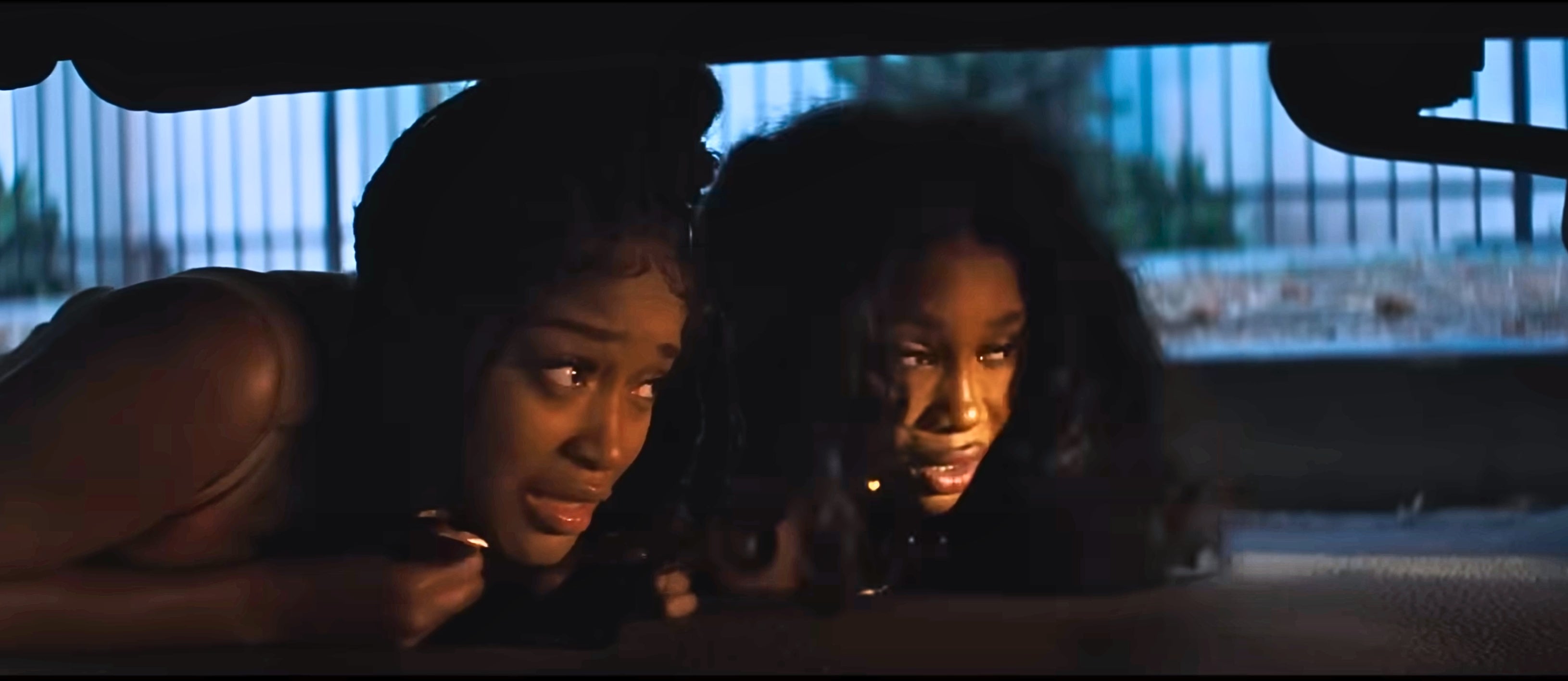 Two people hide under a car, looking intently ahead, suggesting a tense or suspenseful situation