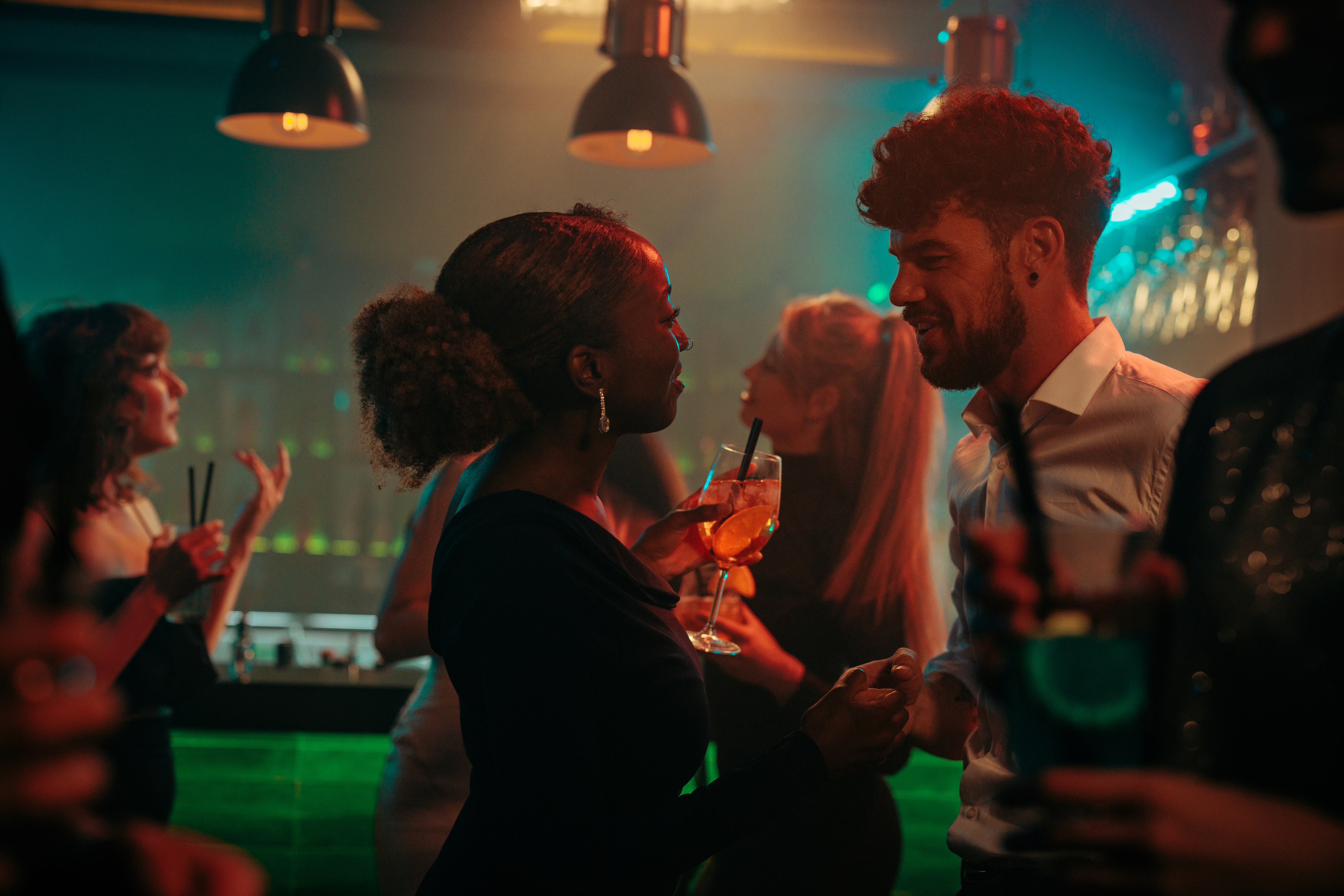 A couple shares a flirtatious moment while holding drinks at a lively nightclub, surrounded by people socializing