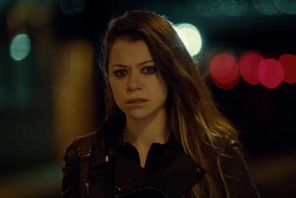 A person with long hair looks serious, standing outdoors at night, wearing a leather jacket