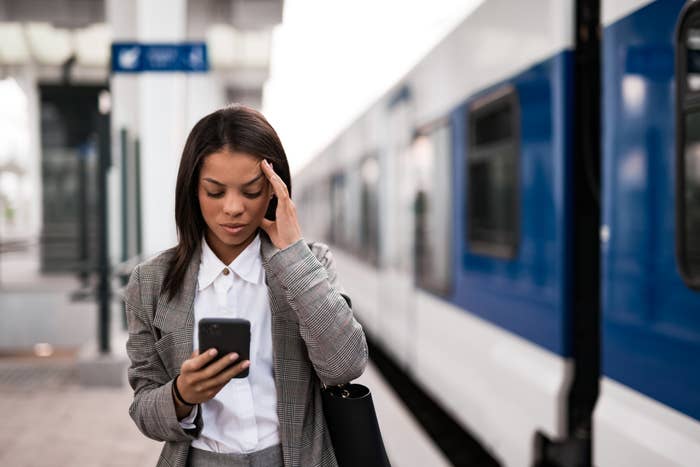 Woman in checkered suit looks at phone, holding head, standing near a train at a station