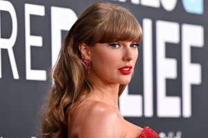 Taylor Swift in elegant gown poses on a red carpet, with long wavy hair and statement jewelry, against a backdrop with partially visible text